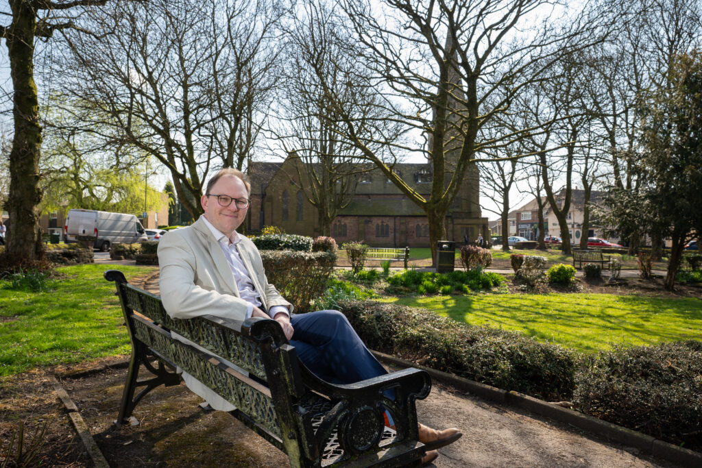 Gareth Snell MP sat on a bench in a park with a Church in the background